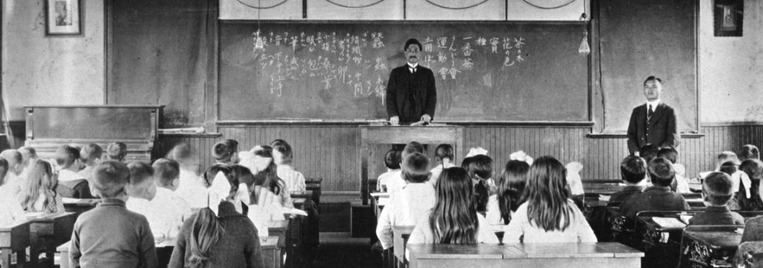 A black and white historical photograph of a classroom filled with children sitting at wooden desks, facing the front of the room. A man in a dark suit stands behind a central podium in front of a large blackboard covered in Japanese kanji and hiragana calligraphy. To his right, another man in a suit stands near the wall. The children, many of whom are girls with large bows in their hair, are seated in organized rows. A piano is visible on the far left, and simple light bulbs hang from the ceiling.