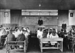 A black and white historical photograph of a classroom filled with children sitting at wooden desks, facing the front of the room. A man in a dark suit stands behind a central podium in front of a large blackboard covered in Japanese kanji and hiragana calligraphy. To his right, another man in a suit stands near the wall. The children, many of whom are girls with large bows in their hair, are seated in organized rows. A piano is visible on the far left, and simple light bulbs hang from the ceiling.