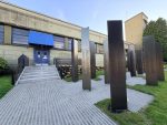 An outdoor memorial with tall dark metal pillars engraved with names stands on a paved stone courtyard outside the Japanese Canadian Hastings Park Interpretive Centre building.