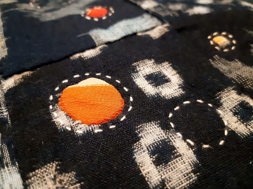 A close-up shot of a textured, black fabric featuring traditional Japanese textile techniques, specifically Sashiko stitching and Boro-inspired patching.