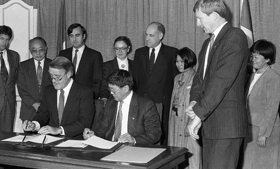 Prime Minister Brian Mulroney and Art Miki sitting down at the signing of the redress. Roger Obata, Audrey Kobayashi, Gerry Weiner, and Maryka Omatsu are among the people standing behind them.

NNMCC Gordon King Collection 2010.32.26