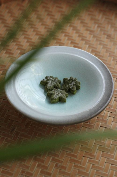 A high-angle, vertical shot shows three small, dark green Japanese sweets (higashi) shaped like maple leaves, resting in the center of a pale blue, shallow ceramic bowl. The sweets are lightly dusted with white powdered sugar. The bowl sits on a textured, light-brown woven bamboo mat. In the foreground and background, blurry green leaves frame the shot, creating a soft, natural depth of field.