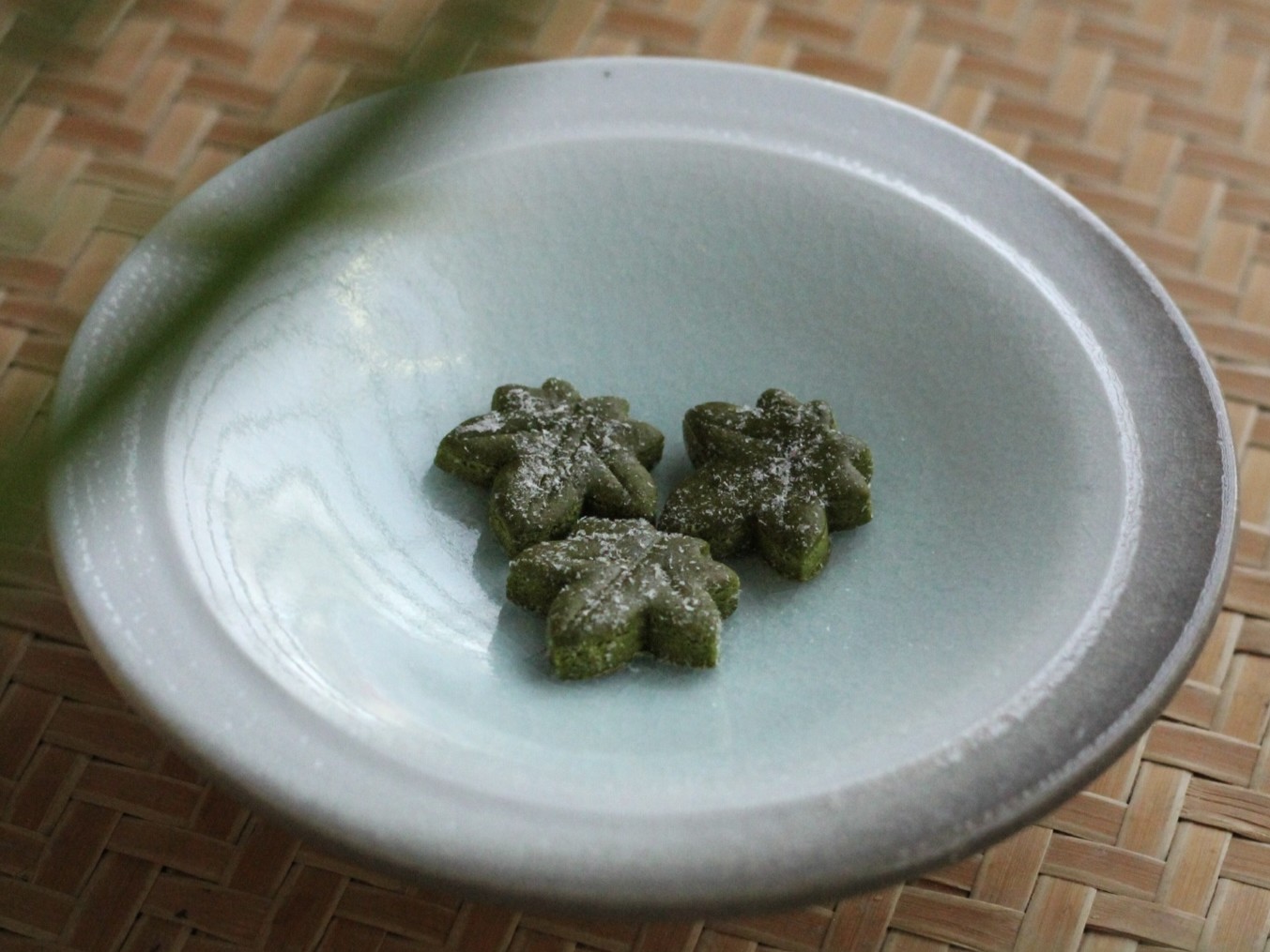 A high-angle, vertical shot shows three small, dark green Japanese sweets (higashi) shaped like maple leaves, resting in the center of a pale blue, shallow ceramic bowl. The sweets are lightly dusted with white powdered sugar. The bowl sits on a textured, light-brown woven bamboo mat. In the foreground and background, blurry green leaves frame the shot, creating a soft, natural depth of field.
