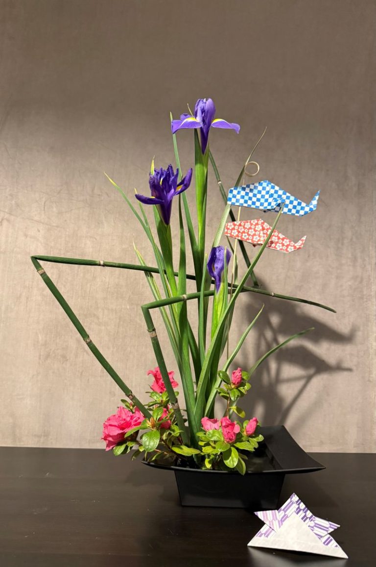 A sogetsu school ikebana flower arrangement with irises and carp origami in a shallow black vase with a simple background. A kabuto origami in the foreground.