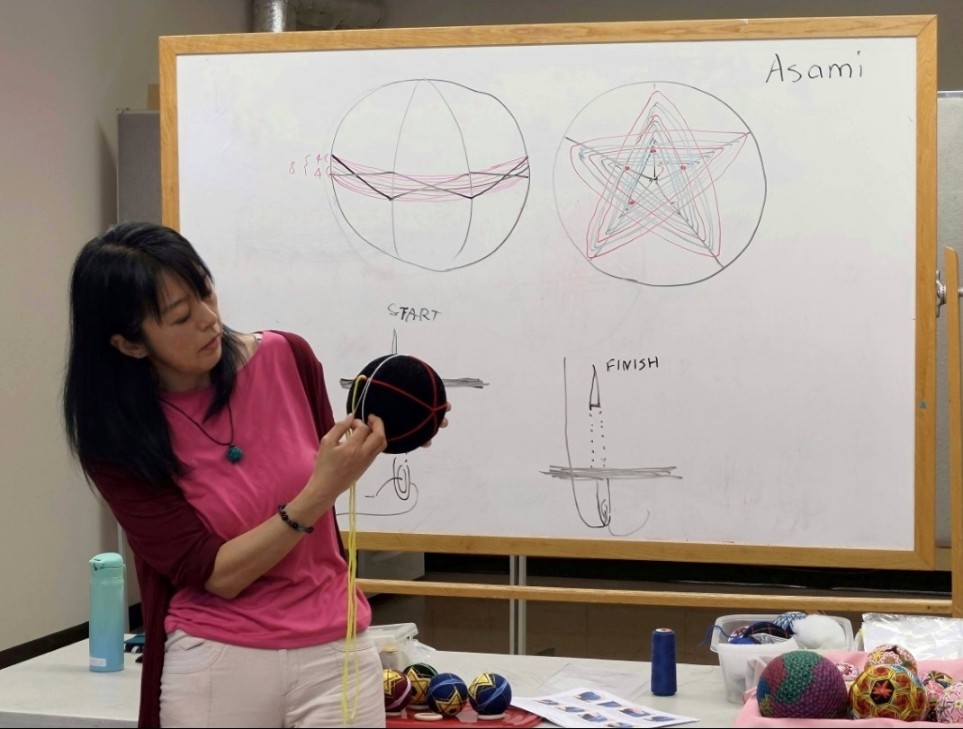 A class room photo of a lady holding a temari in her hand demonstrating where to thread. In the background is a whiteboard with two diagrams of the temari design, as well as drawings that show how to start and finish a stitch.