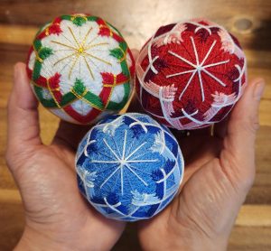 Three colourful temari balls held in two hands. One is made with red, pink and white threads, another with blues, and the third with red and green Christmas colours.