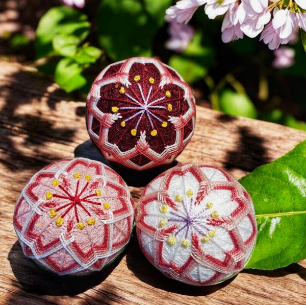Three cherry blossom designed temari balls on a bench outdoors photographed under the sunshine with a branch of cherry blossoms in the corner.
