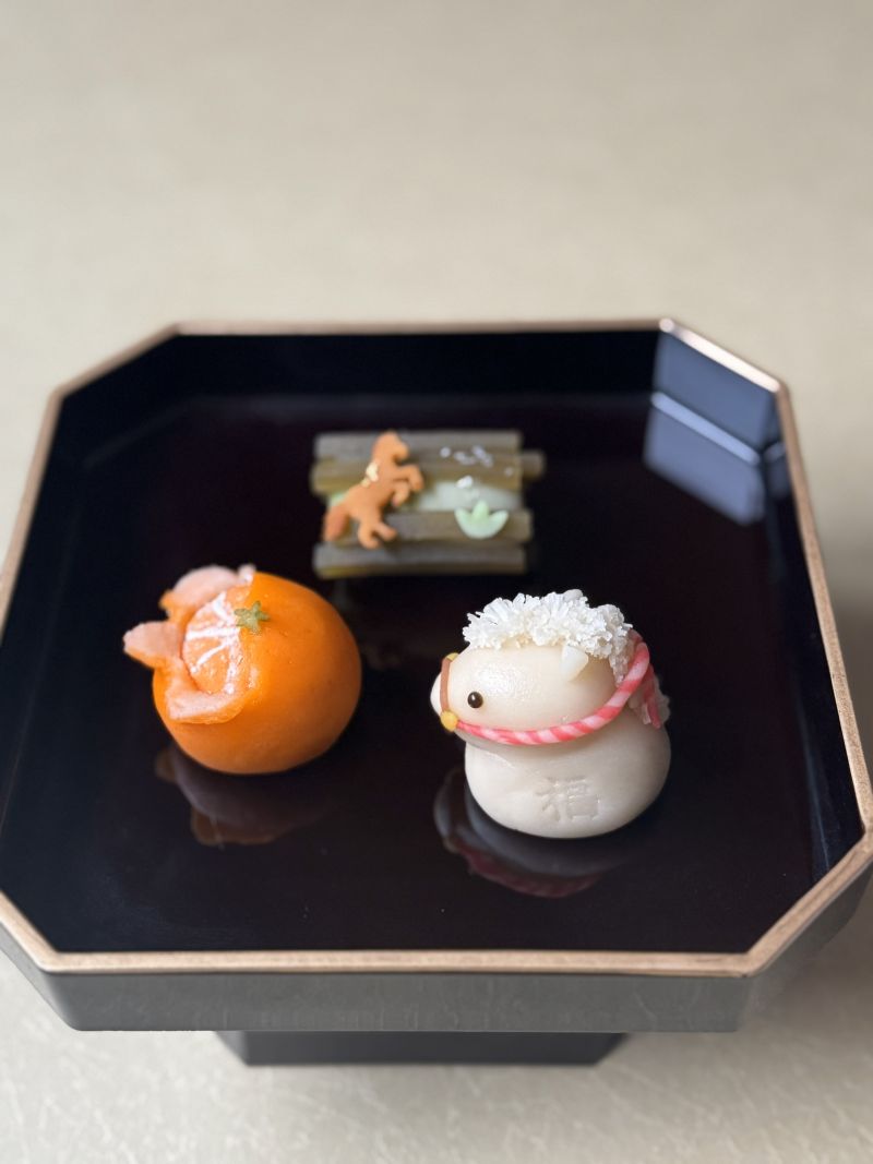 A set of three Japanese New Year wagashi sweets on a black tray: a realistic mandarin orange (mikan), a white horse with the kanji for 'good fortune' (福), and a rectangular sweet featuring a horse silhouette and bamboo.