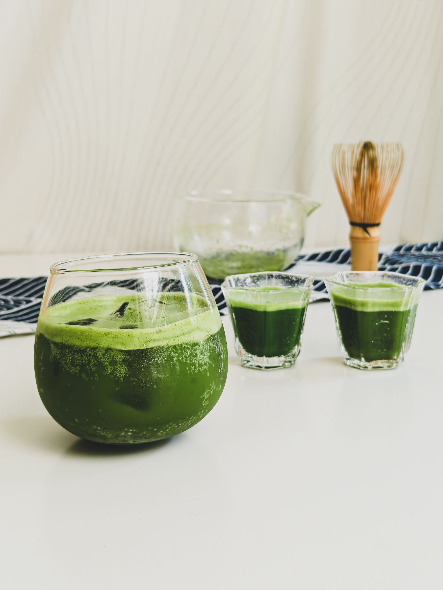Iced matcha in one large and 2 small glasses in the front ground. Tea whisk chasen and empty glass bowl used to whist match blurred in the background on a white and blue tenugui towel.