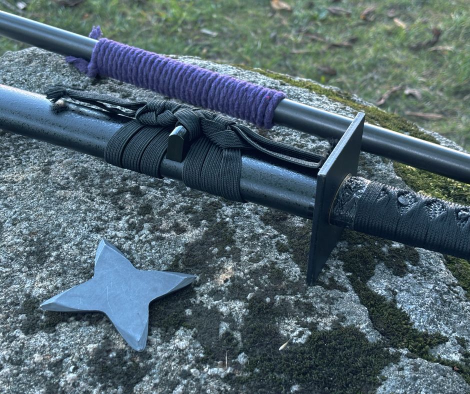 A four-pointed metal shuriken (throwing star) sits on the rock. Behind it lies a katana in its black scabbard (saya), which is intricately wrapped with a black cord (sageo). The sword features a square handguard (tsuba) and a black textured hilt (tsuka). Partially visible in the background is a long, dark staff or weapon handle with a section wrapped in purple cord. The scene is set against a natural, slightly blurred backdrop of grass and fallen leaves.