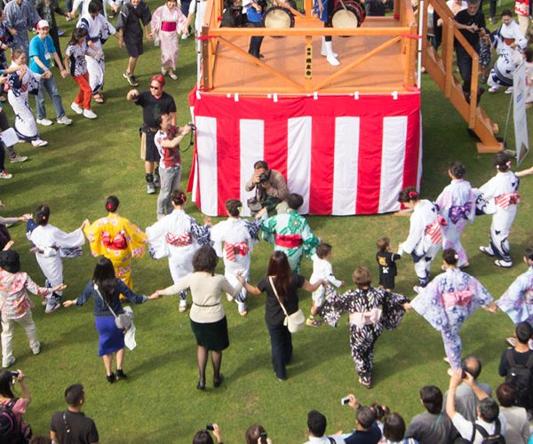 A panoramic aerial view of an outdoor Japanese festival (Matsuri) on a large green lawn. A crowd of people, some in traditional yukata, are dancing in a circle (Bon Odori) around a central wooden yagura stage. The scene is surrounded by festival tents, vendor booths, and many spectators enjoying the sunlit event.