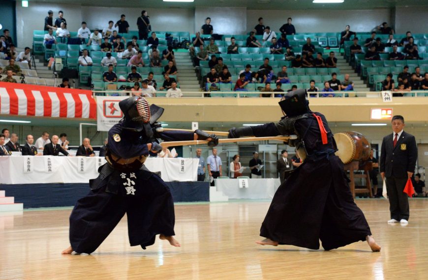 In a large indoor arena, two martial artists dressed in dark blue bogu (protective armor) and hakama engage in a Jukendo match on a polished wooden floor. They are both lunging forward, thrusting long wooden bayonets, known as mokuju, toward one another. The practitioner on the left has "Miyagi" and "Kono" written in white kanji on their tare (waist protector). To the right, a referee in a black suit holds a small red flag, closely monitoring the exchange. In the background, rows of spectators sit in green stadium seating.