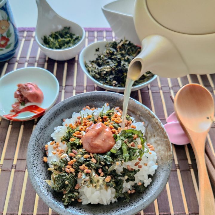 Pouring hot tea from a teapot over a blow of ochazuke with tea leaves. A wooden spoon on the right side, and small plates with ingredients on a table mat.