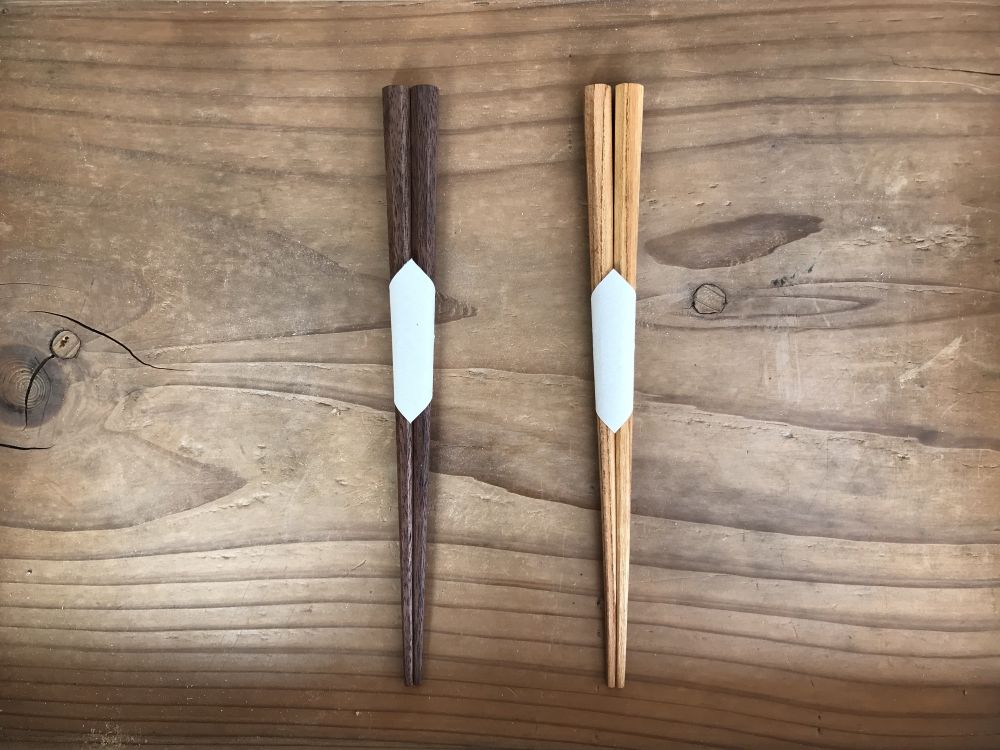 A top-down, minimalist shot showcases two pairs of handcrafted wooden chopsticks (hashi) resting vertically on a rustic, grained wooden surface.