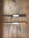 A top-down, minimalist shot showcases two pairs of handcrafted wooden chopsticks (hashi) resting vertically on a rustic, grained wooden surface.