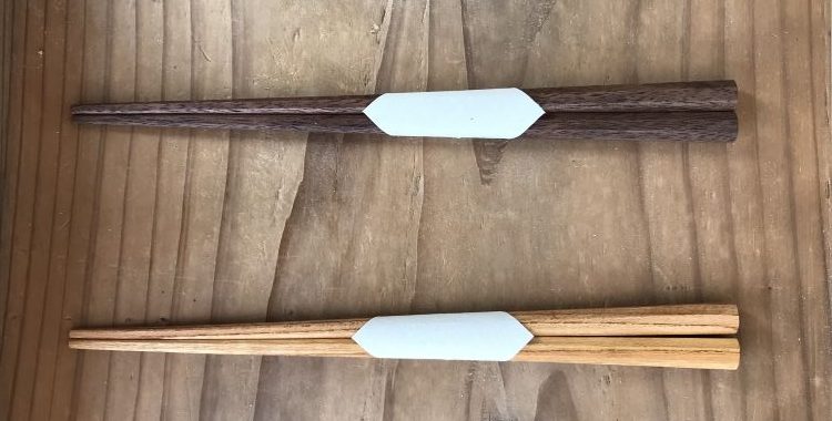 A top-down, minimalist shot showcases two pairs of handcrafted wooden chopsticks (hashi) resting vertically on a rustic, grained wooden surface.