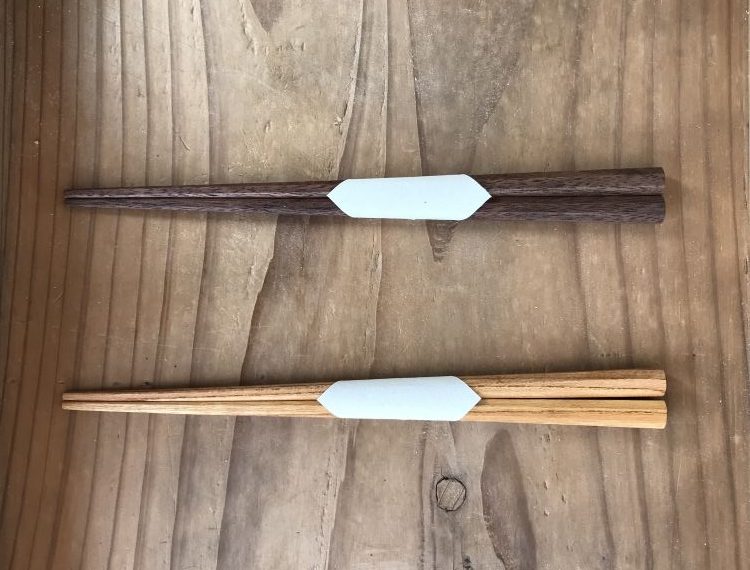A top-down, minimalist shot showcases two pairs of handcrafted wooden chopsticks (hashi) resting vertically on a rustic, grained wooden surface.