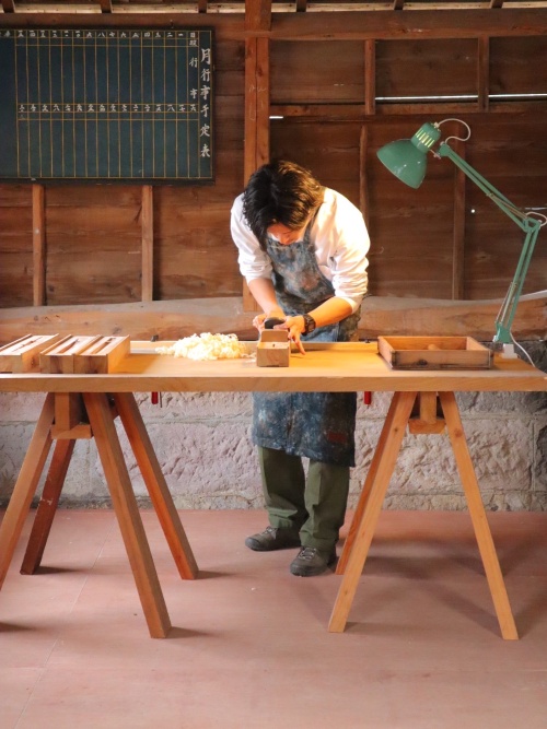 A wide, eye-level shot captures a craftsman at work in a rustic woodworking studio, focused on the traditional art of making chopsticks. The artisan, a man with dark hair wearing a white shirt and a splattered blue apron, leans over a long wooden workbench supported by sawhorses. He is using a traditional Japanese hand plane (kanna) to shave a thin piece of wood, with a small pile of light-colored wood shavings accumulating on the table. To his left are several wooden jigs or molds, and a green architect-style desk lamp is clamped to the right side of the bench, casting light on his workspace. The background features aged wooden walls and a stone foundation. On the wall behind him hangs a dark chalkboard with a grid and Japanese characters, likely a schedule or production chart. The overall mood is one of quiet, diligent craftsmanship within a traditional workshop setting.