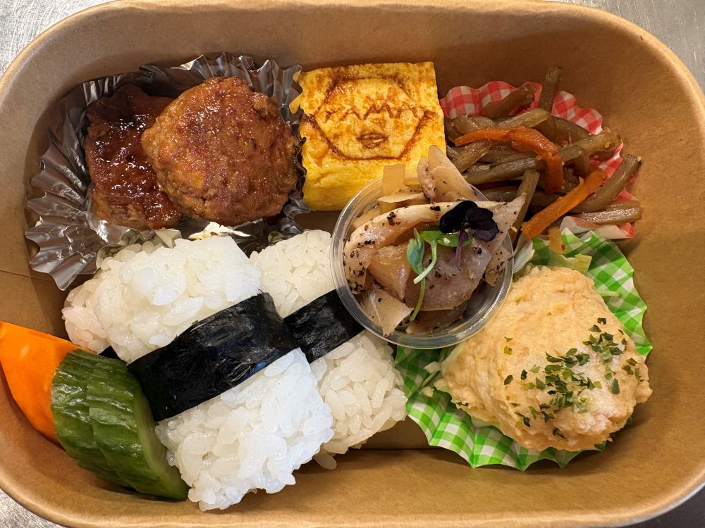 A top-down, brightly lit shot features a meticulously arranged Japanese bento box in a tan cardboard container. The meal is a vibrant assortment of colors and textures: Bottom left: Two rounded mounds of white rice are joined by a crisp band of dark green nori (seaweed), accompanied by slices of bright orange carrot and green pickled cucumber. Top left: Two glazed meatballs or pieces of protein sit in a silver foil cupcake liner. Top center: A square of tamagoyaki (rolled omelet) is branded with a "YAMA" logo and a stylized face. Top right: A small serving of kinpira gobo (braised burdock root and carrot) rests in a red-and-white checkered liner. Bottom right: A scoop of creamy potato salad topped with green herbs sits in a green-and-white checkered liner. Center: A small clear plastic cup contains a seasoned vegetable or seafood salad garnished with a tiny purple flower and a green sprout.