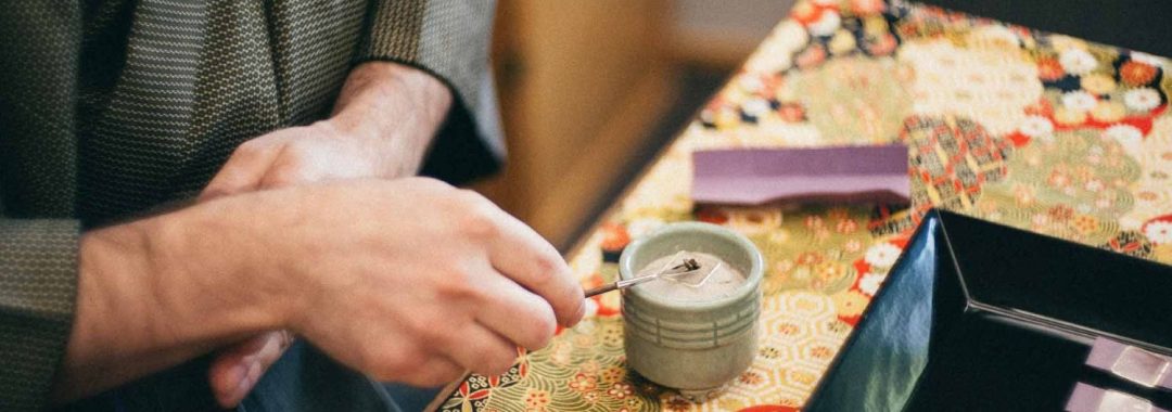A close-up, slightly high-angle shot captures a person participating in Kōdō, the traditional Japanese art of appreciating incense. The person, wearing a dark olive-green patterned kimono, is shown from the chest down. Their hands are the focus as they use a thin metal tool to meticulously arrange white ash inside a small, pale green ceramic incense burner (koro). The scene is set on a table covered with a vibrant, multi-colored fabric featuring traditional Japanese floral and geometric patterns in shades of red, gold, green, and black. To the right of the burner sits a square black lacquer tray containing small purple slips of paper, and a single purple tool rest sits behind the burner. The background is softly blurred, showing the warm wood and paper of a traditional shoji screen, creating a quiet and contemplative atmosphere.