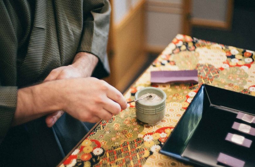 A close-up, slightly high-angle shot captures a person participating in Kōdō, the traditional Japanese art of appreciating incense. The person, wearing a dark olive-green patterned kimono, is shown from the chest down. Their hands are the focus as they use a thin metal tool to meticulously arrange white ash inside a small, pale green ceramic incense burner (koro). The scene is set on a table covered with a vibrant, multi-colored fabric featuring traditional Japanese floral and geometric patterns in shades of red, gold, green, and black. To the right of the burner sits a square black lacquer tray containing small purple slips of paper, and a single purple tool rest sits behind the burner. The background is softly blurred, showing the warm wood and paper of a traditional shoji screen, creating a quiet and contemplative atmosphere.