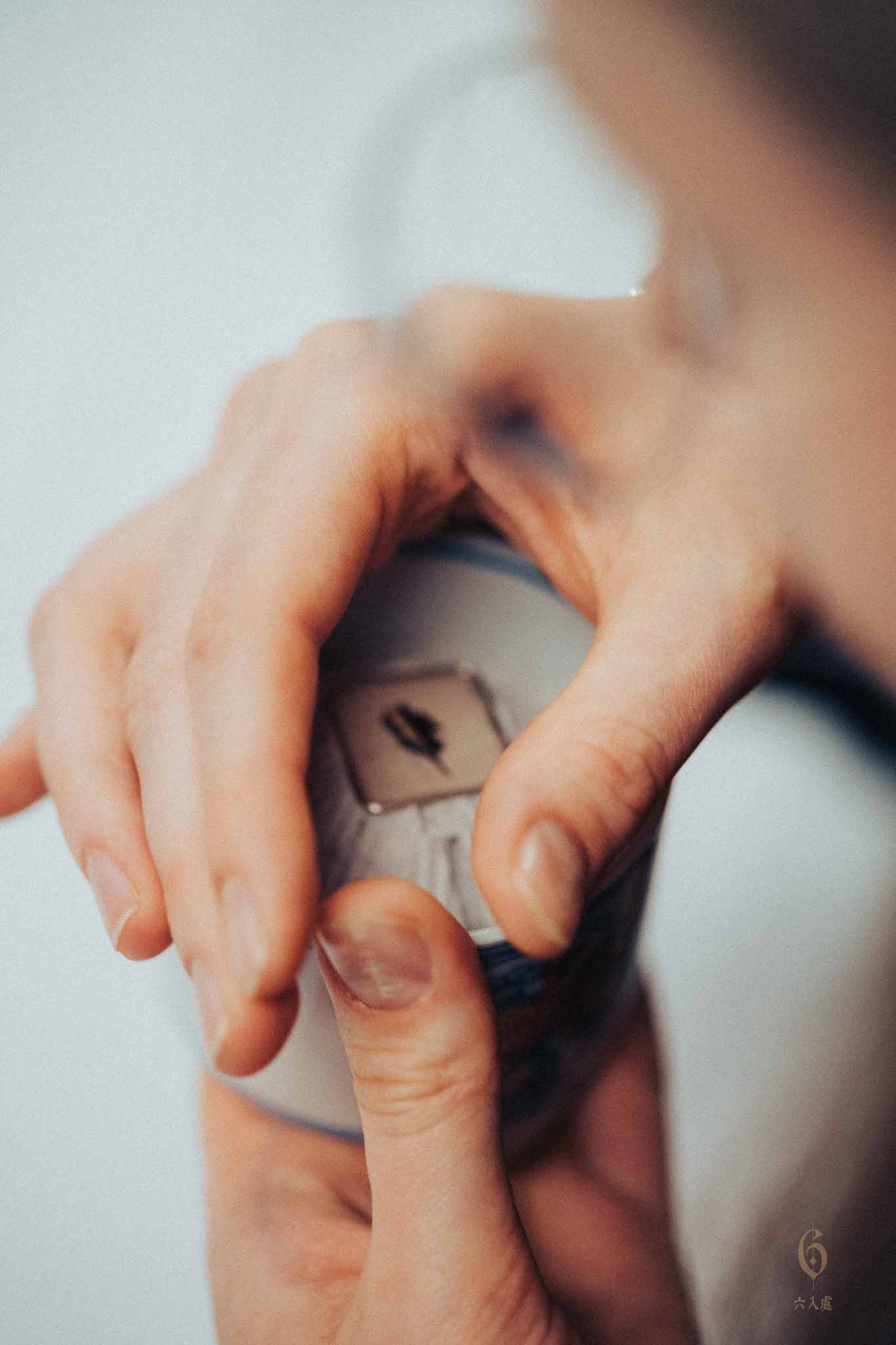 An extreme close-up, high-angle shot captures the intimate moment of a participant "listening" to incense during a Kōdō ceremony. The person's hands are cupped delicately over the top of a ceramic incense burner (koro), creating a small chimney with their thumb and forefinger to direct the fragrant smoke toward their nose. The top of the burner reveals meticulously raked white ash with a small square of mica (gin-yami) resting in the center, upon which a tiny piece of aromatic wood is heated. The lighting is soft and diffused, focusing on the texture of the skin and the precise positioning of the fingers. The background is a minimalist, out-of-focus white, emphasizing the quiet concentration and sensory experience of the ritual. A small, subtle logo is visible in the bottom right corner.