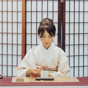 A wide, eye-level shot captures a woman performing a Kōdō (incense ceremony) with graceful precision. She is seated behind a table covered in a white cloth with a maroon runner. Dressed in an elegant, off-white kimono with a delicate floral motif, she looks down with a calm, focused expression as she uses a small tool to tend to a white ceramic incense burner. On the table, several traditional tools are neatly arranged, including a small red vase holding thin metal instruments used for manipulating the ash and incense. The background is dominated by a large, traditional Japanese shoji screen made of dark wood and translucent paper panels, which creates a structured and serene backdrop. To the far left, a vertical scroll with Japanese calligraphy adds to the cultural authenticity of the setting. The lighting is soft and even, highlighting the meditative and refined nature of the ritual.