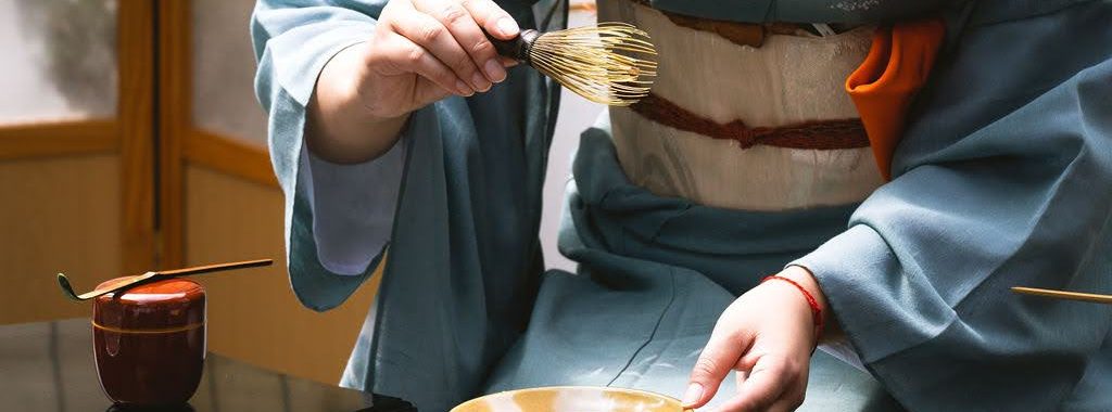 A close-up, eye-level shot focuses on the precise movements of a tea practitioner during a traditional Japanese tea ceremony. The practitioner, wearing a blue-grey kimono with a white and tan obi, is shown from the torso down. Her right hand delicately holds a bamboo tea whisk (chasen) over a shallow, golden-brown ceramic tea bowl (chawan) that she stabilizes with her left hand. To the left, a small, dark red lacquer tea caddy (natsume) sits on the polished black table, with a bamboo tea scoop (chashaku) resting across its lid. The background is softly blurred, showing the warm tones of a shoji screen and a reed mat, which maintains the focus on the ritualistic preparation. The lighting is bright and crisp, highlighting the textures of the bamboo whisk and the fine silk of the kimono, capturing a moment of quiet, intentional grace.