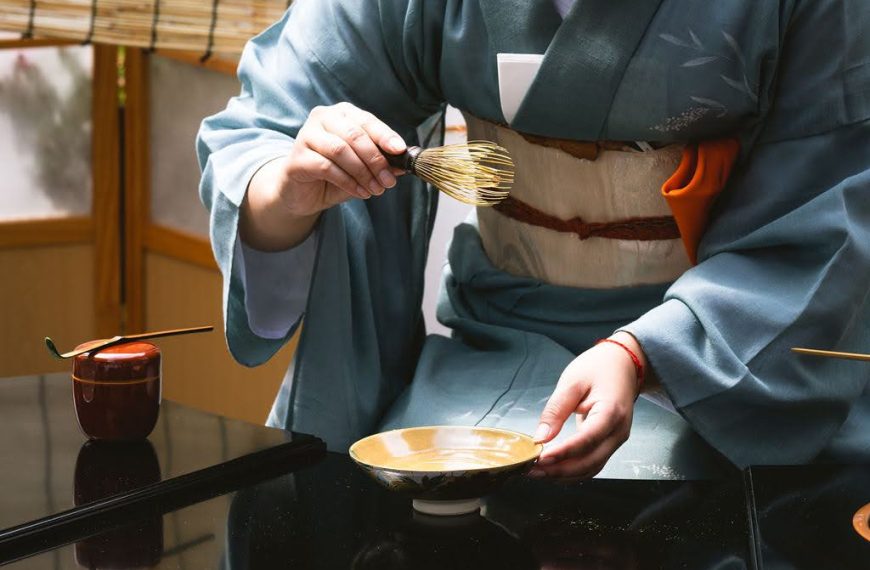 A close-up, eye-level shot focuses on the precise movements of a tea practitioner during a traditional Japanese tea ceremony. The practitioner, wearing a blue-grey kimono with a white and tan obi, is shown from the torso down. Her right hand delicately holds a bamboo tea whisk (chasen) over a shallow, golden-brown ceramic tea bowl (chawan) that she stabilizes with her left hand. To the left, a small, dark red lacquer tea caddy (natsume) sits on the polished black table, with a bamboo tea scoop (chashaku) resting across its lid. The background is softly blurred, showing the warm tones of a shoji screen and a reed mat, which maintains the focus on the ritualistic preparation. The lighting is bright and crisp, highlighting the textures of the bamboo whisk and the fine silk of the kimono, capturing a moment of quiet, intentional grace.