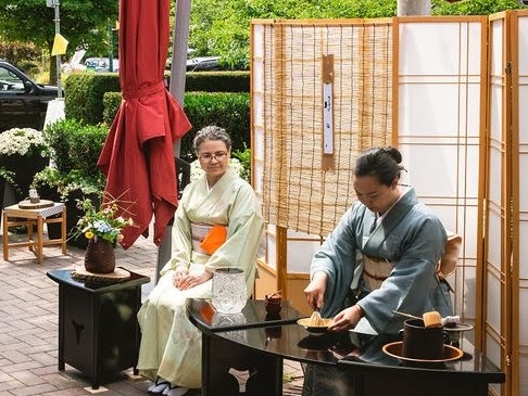 A high-angle, eye-level shot captures a traditional Japanese tea ceremony taking place outdoors on a brick patio under a large red umbrella. Two women, both in their late 40s or early 50s, are dressed in traditional kimonos. On the right, a woman in a blue-grey kimono is seated behind a black, curved wooden table, meticulously whisking matcha in a small bowl. Her dark hair is pulled back, and she is focused on the task. On the table, there are several tea-related items, including a wooden whisk, a small container, and a larger dark pot. To her left, another woman in a pale green kimono with an orange obi sash sits calmly, watching the ceremony. She has greyish-brown hair and wears glasses. Behind them, a tall, three-paneled shoji screen provides a backdrop, with a reed mat hanging in the center. The surrounding area features lush green trees and bushes