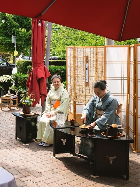 A high-angle, eye-level shot captures a traditional Japanese tea ceremony taking place outdoors on a brick patio under a large red umbrella. Two women, both in their late 40s or early 50s, are dressed in traditional kimonos. On the right, a woman in a blue-grey kimono is seated behind a black, curved wooden table, meticulously whisking matcha in a small bowl. Her dark hair is pulled back, and she is focused on the task. On the table, there are several tea-related items, including a wooden whisk, a small container, and a larger dark pot. To her left, another woman in a pale green kimono with an orange obi sash sits calmly, watching the ceremony. She has greyish-brown hair and wears glasses. Behind them, a tall, three-paneled shoji screen provides a backdrop, with a reed mat hanging in the center. The surrounding area features lush green trees and bushes