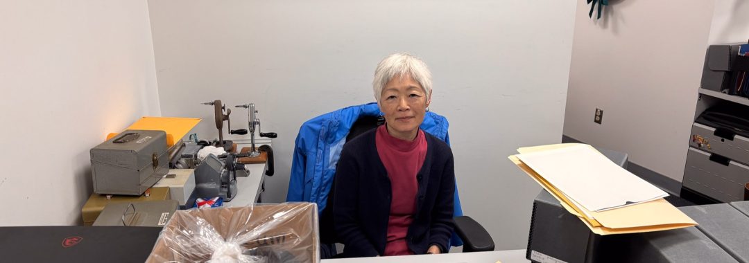 A volunteer with short grey hair, Joy, sits at a desk in the museum's archives department. She is wearing a pink shirt and a navy cardigan, with a blue jacket draped over her chair. Her workspace includes a laptop, archival storage boxes, and various specialized equipment for handling historical materials.