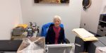 A volunteer with short grey hair, Joy, sits at a desk in the museum's archives department. She is wearing a pink shirt and a navy cardigan, with a blue jacket draped over her chair. Her workspace includes a laptop, archival storage boxes, and various specialized equipment for handling historical materials.