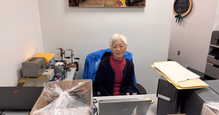 A volunteer with short grey hair, Joy, sits at a desk in the museum's archives department. She is wearing a pink shirt and a navy cardigan, with a blue jacket draped over her chair. Her workspace includes a laptop, archival storage boxes, and various specialized equipment for handling historical materials.
