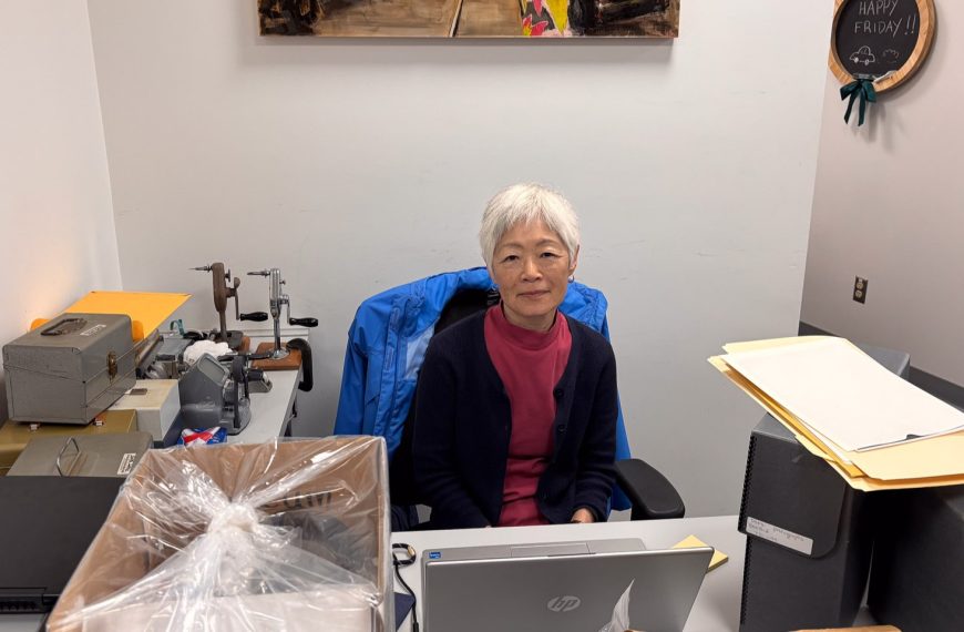 A volunteer with short grey hair, Joy, sits at a desk in the museum's archives department. She is wearing a pink shirt and a navy cardigan, with a blue jacket draped over her chair. Her workspace includes a laptop, archival storage boxes, and various specialized equipment for handling historical materials.