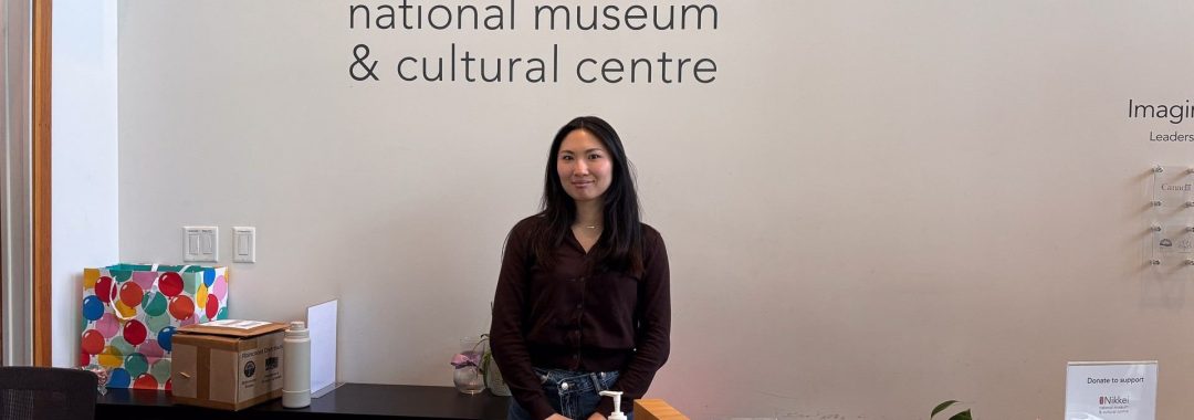 A smiling woman, Nona, stands behind the wooden reception desk at the Nikkei National Museum & Cultural Centre. She is wearing a dark brown cardigan and jeans. Behind her on the white wall is the large black and red logo of the museum. The desk features informational brochures, greeting cards, and a donation box.