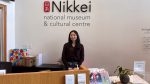A smiling woman, Nona, stands behind the wooden reception desk at the Nikkei National Museum & Cultural Centre. She is wearing a dark brown cardigan and jeans. Behind her on the white wall is the large black and red logo of the museum. The desk features informational brochures, greeting cards, and a donation box.