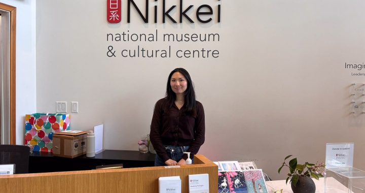 A smiling woman, Nona, stands behind the wooden reception desk at the Nikkei National Museum & Cultural Centre. She is wearing a dark brown cardigan and jeans. Behind her on the white wall is the large black and red logo of the museum. The desk features informational brochures, greeting cards, and a donation box.