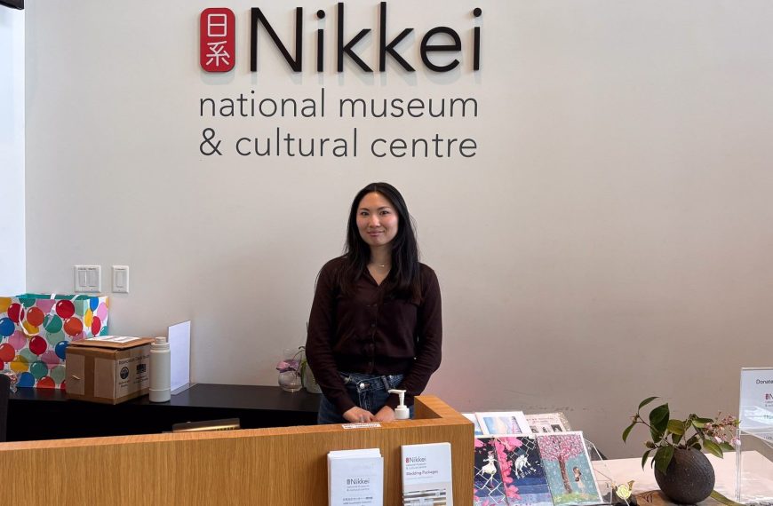 A smiling woman, Nona, stands behind the wooden reception desk at the Nikkei National Museum & Cultural Centre. She is wearing a dark brown cardigan and jeans. Behind her on the white wall is the large black and red logo of the museum. The desk features informational brochures, greeting cards, and a donation box.