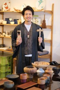 A medium-shot, eye-level photograph shows a smiling Japanese man standing behind a table of handcrafted wooden and ceramic items in what appears to be a shop or studio. He has short, dark, styled hair and a light goatee, wearing a dark grey haori over a patterned brown vest and a white t-shirt. He holds a wooden hand plane (kanna) in his right hand and a wooden mallet in his left.