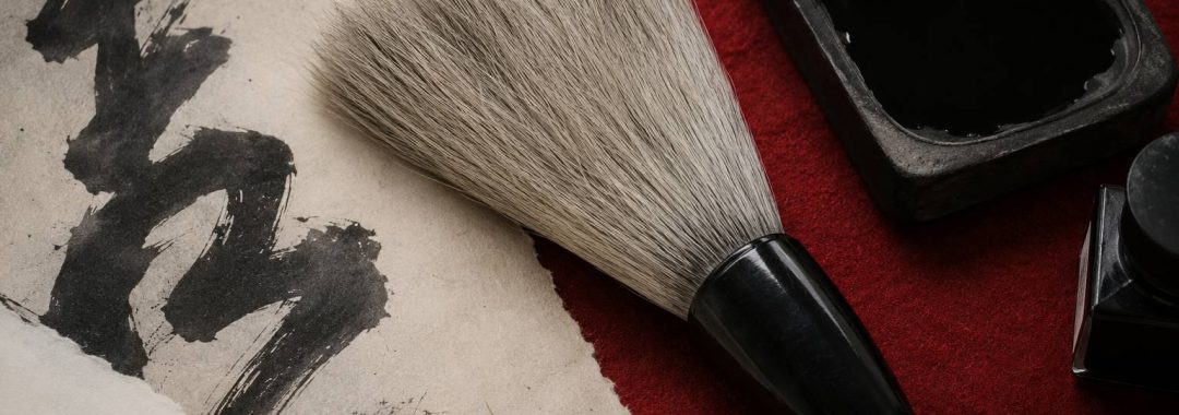 A close-up shot of Japanese calligraphy tools on a red mat, featuring a large brush, an inkstone filled with black ink, and a piece of textured paper with a bold calligraphy stroke.