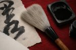 A close-up shot of Japanese calligraphy tools on a red mat, featuring a large brush, an inkstone filled with black ink, and a piece of textured paper with a bold calligraphy stroke.