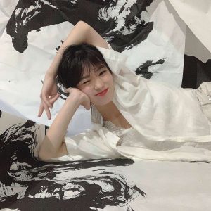 A young woman with short dark hair smiling and winking while reclining amidst large sheets of black and white abstract ink calligraphy art.