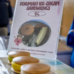 A row of four dorayaki ice-cream sandwiches with colorful fillings displayed in front of a fundraising sign that reads "Dorayaki Ice-Cream Sandwiches $6."