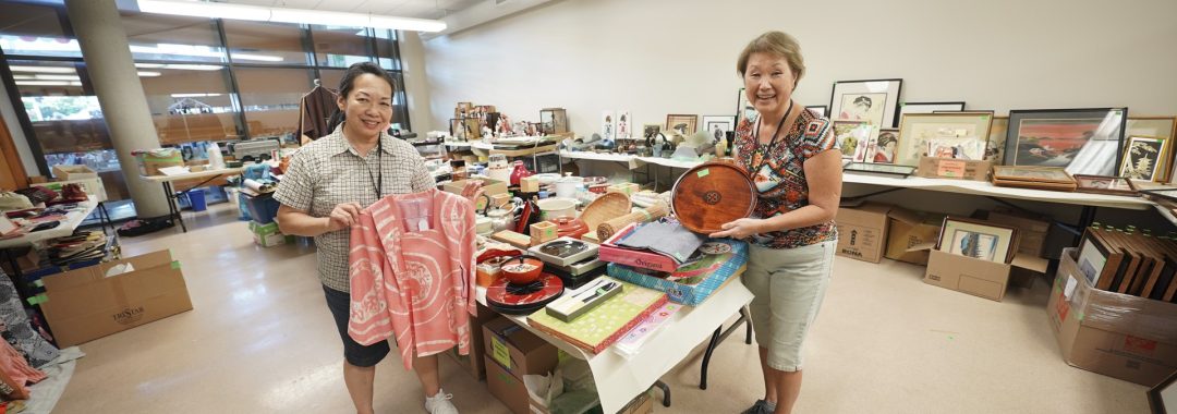 Two smiling volunteers holding a pink kimono jacket and wooden tray in a room filled with donated Japanese items.