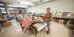Two smiling volunteers holding a pink kimono jacket and wooden tray in a room filled with donated Japanese items.