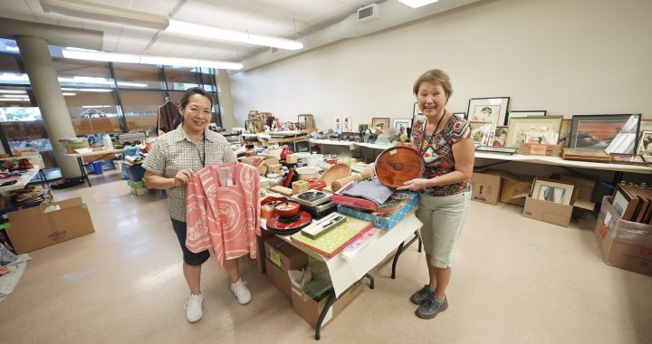 Two smiling volunteers holding a pink kimono jacket and wooden tray in a room filled with donated Japanese items.