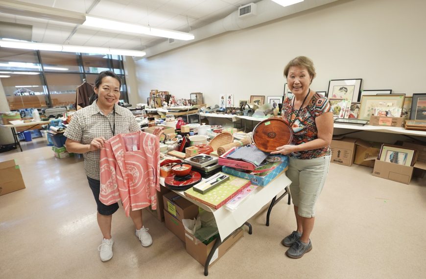 Two smiling volunteers holding a pink kimono jacket and wooden tray in a room filled with donated Japanese items.
