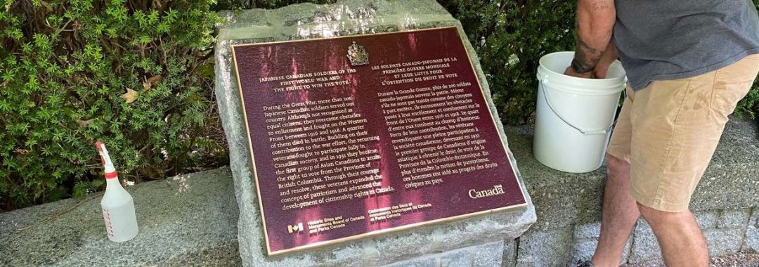 A man leaning down to wash a bronze Japanese Canadian Soldiers historical plaque set in stone outdoors.