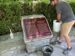 A man leaning down to wash a bronze Japanese Canadian Soldiers historical plaque set in stone outdoors.